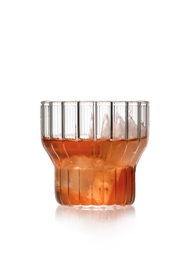 A low, fluted, modern drinking glass filled with ice cubes and amber drink.