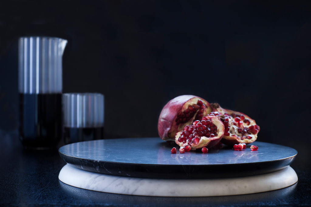 A marble serving board black marble face up with an open pomegranate  placed on it, photographed on a black background, with styled glassware out of focus in the background