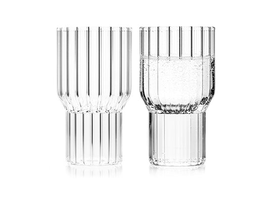 A pair of fluted, contemporary, clear glasses by designer, Felicia Ferrone. One glass is filled with carbonated water, the other is empty. 