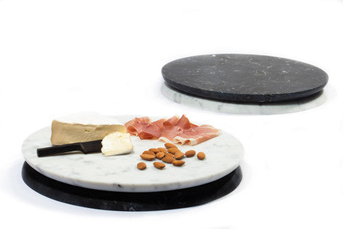 A marble serving board white marble face up with a piece of cheese, prosciutto, and nuts placed on it, photographed on a white background, with another marble serving board with the reverse side up, made of black marble.